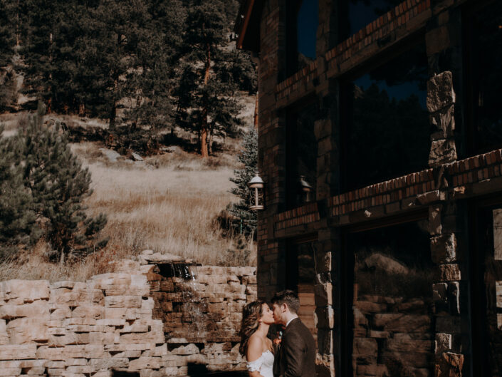 bride and groom kiss in front of stone building and mountain