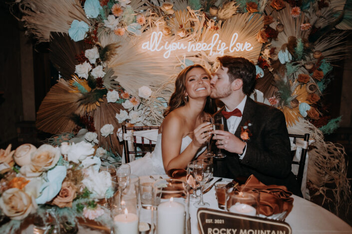 groom kisses the bride's drink during a cheers in front of their floral installation at their dinner table