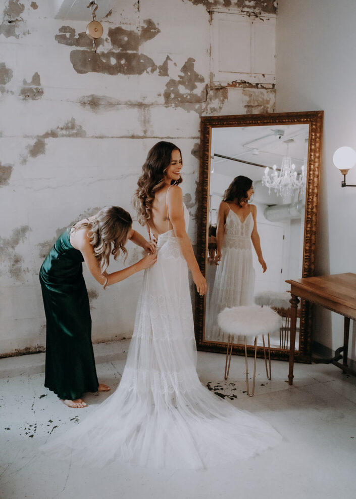 brides sister buttons up her dress in front of mirror