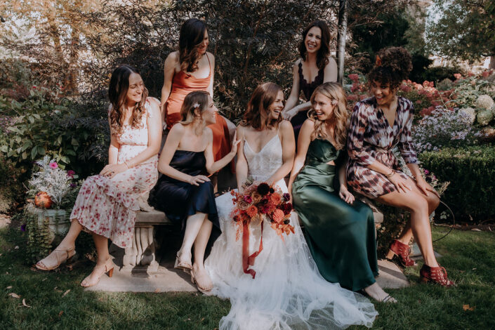 bride and bridesmaid sit on stone bench and laugh together