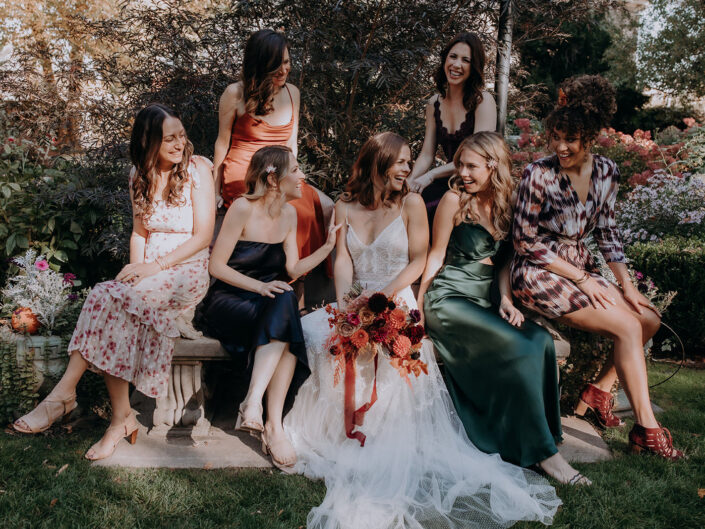bride and bridesmaid sit on stone bench and laugh together